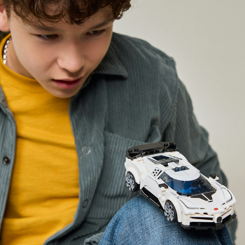 Young child wearing yellow shirt and grey jacket examining white LEGO Bugatti Centodieci hypercar model with blue interior details and black roof spoiler, displayed on denim jeans against neutral background
