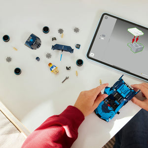 Child in red sleeve assembling blue LEGO Bugatti Vision GT sports car with scattered components including wheels, engine parts, and instruction tablet displayed on white surface