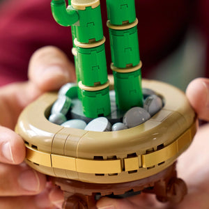 LEGO Lucky Bamboo set 10344 held in hands showing green segmented bamboo stalks with tan bands and beige banana-shaped base containing gray and white LEGO elements against pink blurred background