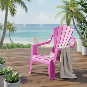 Bright fuchsia pink plastic Adirondack-style children's mini chair on wooden deck with beach and palm trees in background, featuring integrated cup holder and grey knit blanket draped over armrest