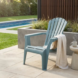 Modern grey-blue Adirondack-style garden chair on paved patio beside swimming pool, with white towel draped over arm, concrete planter with ornamental grasses, and cream side table with mug