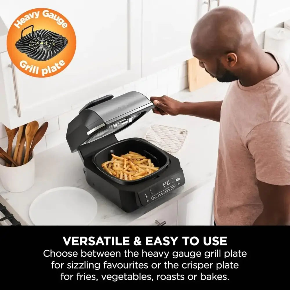 Man using black Ninja 5-in-1 grill and air fryer on white kitchen counter with french fries cooking inside, heavy gauge grill plate feature highlighted in orange circle logo, white plate and wooden utensils beside appliance