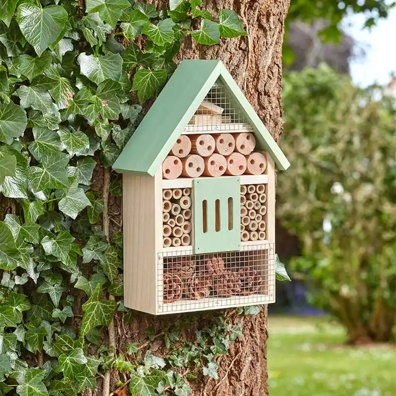 Wooden bug and bee habitat house mounted on tree trunk with sage green roof and door, featuring natural wood sections filled with bamboo tubes and mesh panels, surrounded by green ivy leaves