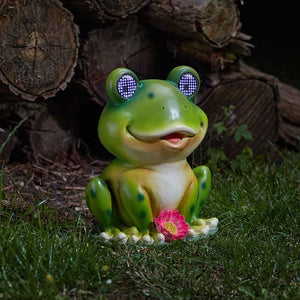 Green garden frog solar light with illuminated LED eyes sitting on moss and grass next to a small pink flower and weathered logs