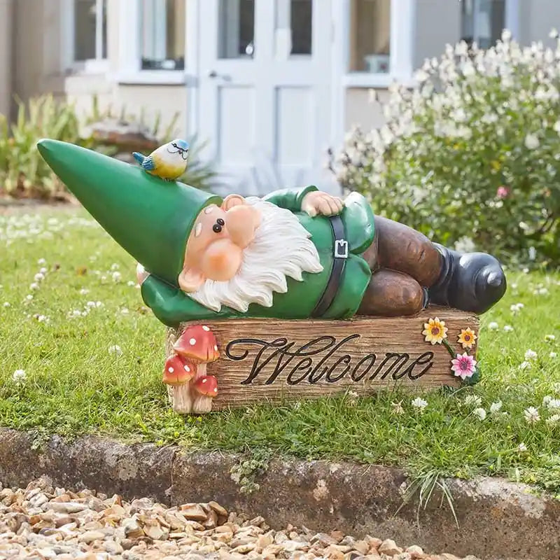 Garden gnome ornament named Wilf wearing green outfit lying down on wooden welcome sign with mushrooms and flowers, blue bird perched on hat, positioned in grass border with white house and hedges in background