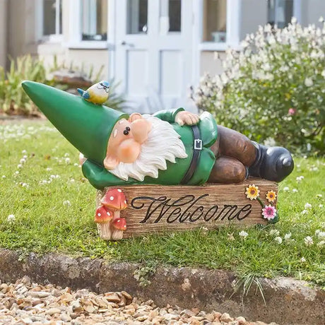 Garden gnome ornament named Wilf wearing green outfit lying down on wooden welcome sign with mushrooms and flowers, blue bird perched on hat, positioned in grass border with white house and hedges in background