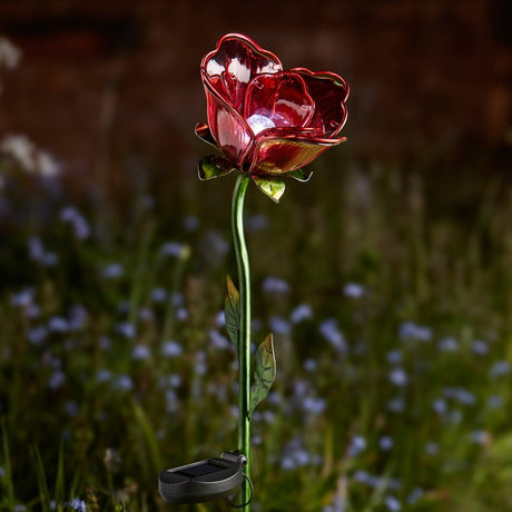 Smart Solar Garden Stake Light in the shape of a red rose with green stem and leaves, illuminated against a blurred garden background with small blue flowers
