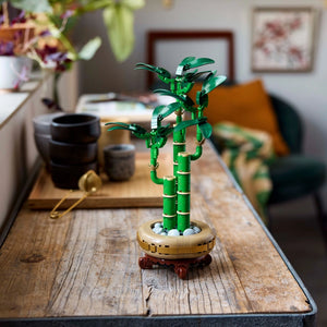 LEGO Lucky Bamboo 10344 set displayed on wooden table featuring green segmented bamboo stalks with leaf elements, tan and brown circular base with decorative stones, positioned in home interior with black ceramic vessels, purple flowers, and framed artwork in background