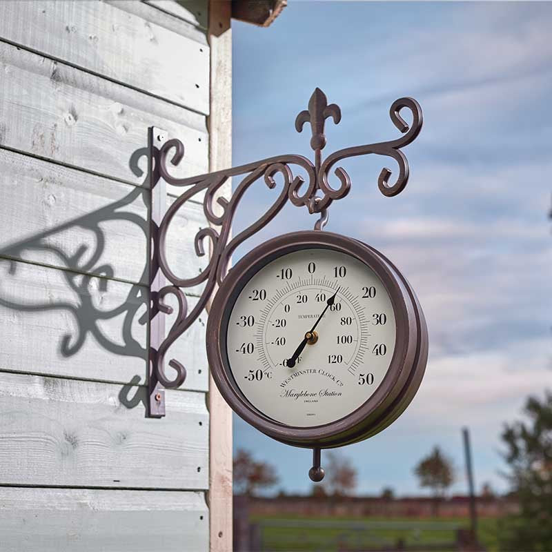Ornate wrought iron garden wall clock and thermometer with cream dial mounted on white wooden house wall against blue sky, displaying temperature scale in Celsius and Fahrenheit