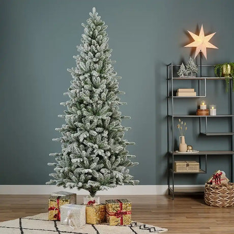 Everlands Snowy Killington Fir 7.5ft pre-lit Christmas tree with frosted white branches and green needles displayed in a contemporary living room with wrapped gifts, gold star topper, industrial shelving unit, and festive decorations on wooden flooring against a sage green wall