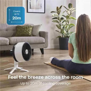 Dimplex Duocool 2 in 1 pedestal desk fan in graphite with white base positioned in a modern living room, demonstrating 20m cooling coverage with woman sitting on yoga mat, featuring gray sofa with green cushions and potted plant in background
