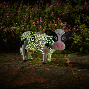 Decorative garden silhouette of a whimsical cow with lime green and black patterned body, white legs and face, pink nose, and a white daisy flower on its side, illuminated at night against a backdrop of pink flowers and green foliage