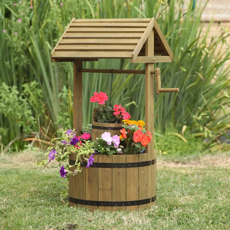 Wooden wishing well planter with shingled roof and crank handle, filled with colorful flowers including red geraniums, pink petunias, yellow marigolds and purple petunias, standing in a garden setting with ornamental grasses in background