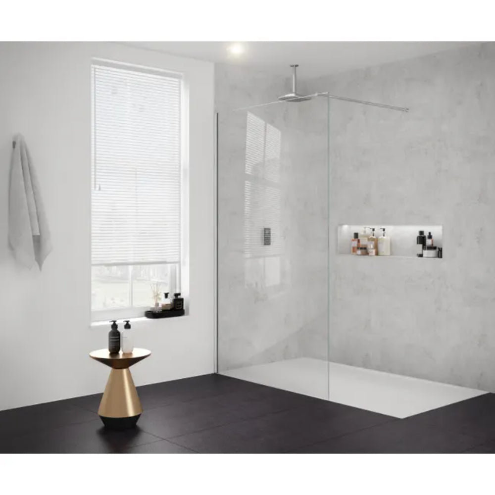 Modern bathroom featuring Merlyn 8mm glass wetroom panel with brushed nickel finish, concrete grey tile walls, black floor tiles, rainfall showerhead, white shower tray, built-in wall niches with toiletries, brass cone side table, white towel rail, and window with horizontal blinds