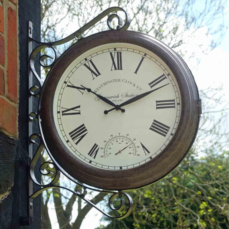 Vintage-style outdoor wall clock with ornate bronze scrollwork bracket, cream face with Roman numerals, and integrated thermometer, mounted on dark wooden post with brick wall and trees visible in background