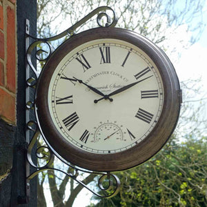 Vintage-style outdoor wall clock with ornate bronze scrollwork bracket, cream face with Roman numerals, and integrated thermometer, mounted on dark wooden post with brick wall and trees visible in background