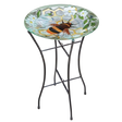 Glass bird bath with painted bee design on round top, supported by black metal stand with curved legs, featuring white flowers and green foliage, ideal for garden decoration