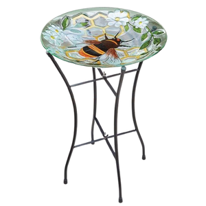 Glass bird bath with painted bee design on round top, supported by black metal stand with curved legs, featuring white flowers and green foliage, ideal for garden decoration
