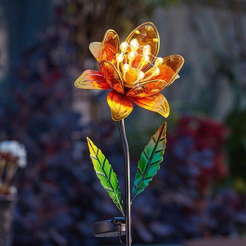 Solar-powered garden stake light with amber and golden yellow glass flower petals, green translucent leaves, and illuminated LED center, displayed in a garden setting