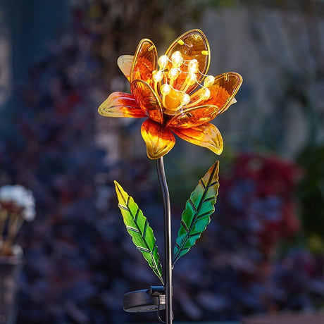 Solar-powered garden stake light with amber and golden yellow glass flower petals, green translucent leaves, and illuminated LED center, displayed in a garden setting