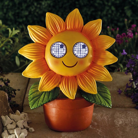 Solar-powered decorative sunflower ornament with yellow and orange petals, smiling face with LED light-up eyes, green leaves, and terracotta pot displayed in a garden setting with purple flowers and stones