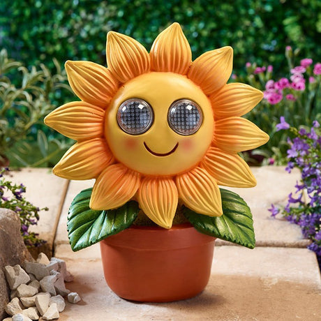 Cheerful yellow and orange solar-powered sunflower ornament with large round eyes in terracotta pot, displayed in garden setting with purple flowers and green foliage
