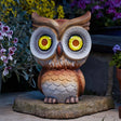 Decorative wooden owl garden statue with glowing yellow and red solar-powered LED eyes, perched on a stone base surrounded by purple and pink flowering plants in a garden setting