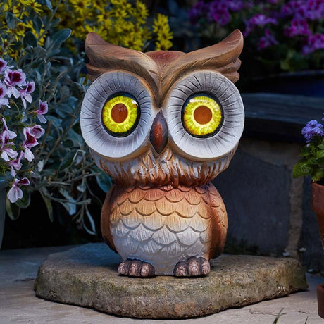 Decorative wooden owl garden statue with glowing yellow and red solar-powered LED eyes, perched on a stone base surrounded by purple and pink flowering plants in a garden setting