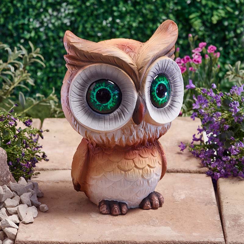 Decorative ceramic owl garden statue with large turquoise green eyes and brown plumage, positioned on a stone pathway surrounded by purple flowers and green foliage, solar-powered outdoor lighting ornament