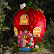 Smart Solar Garden Strawberry House ornament with bright red body, yellow-lit windows, pink door, green leaf top and decorative roses, displayed on moss-covered base in garden setting