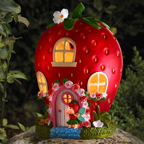 Smart Solar Garden Strawberry House ornament with bright red body, yellow-lit windows, pink door, green leaf top and decorative roses, displayed on moss-covered base in garden setting