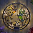 Solar-powered garden ornament featuring two illuminated hummingbirds with colored glass wings in amber and green, surrounded by decorative black metal flowers and foliage on a circular brass-toned metal disc, mounted on wooden fence with purple and red flowers visible