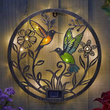 Solar-powered garden ornament featuring two illuminated hummingbirds with colored glass wings in amber and green, surrounded by decorative black metal flowers and foliage on a circular brass-toned metal disc, mounted on wooden fence with purple and red flowers visible