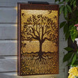 Solar powered metal garden panel with tree of life design, featuring bronze frame with golden backlit silhouette of tree and intricate root pattern, mounted on wooden wall with climbing ivy and yellow flowers
