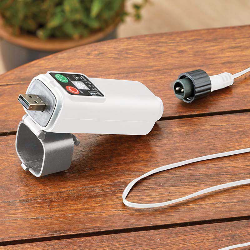 Smart Garden rechargeable garden string lights controller with white casing and black wrist band attachment sitting on wooden decking, showing green and red control buttons on digital display, with white cable and connector visible