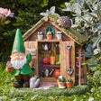 Garden gnome figurine with green pointed hat standing beside a wooden shed miniature with moss-covered roof, featuring tiny potted flowers, gardening tools, watering can and decorative plants in a natural garden setting