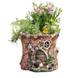 A whimsical fairy garden planter shaped like a tree trunk with a small door, window, hedgehog figures, and green plants with pink and purple flowers, shown on a plain white background.