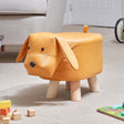 Tan leather dog-shaped footstool with floppy ears, black button eyes, and natural wooden legs, positioned on a beige carpet in a living room setting with toys nearby