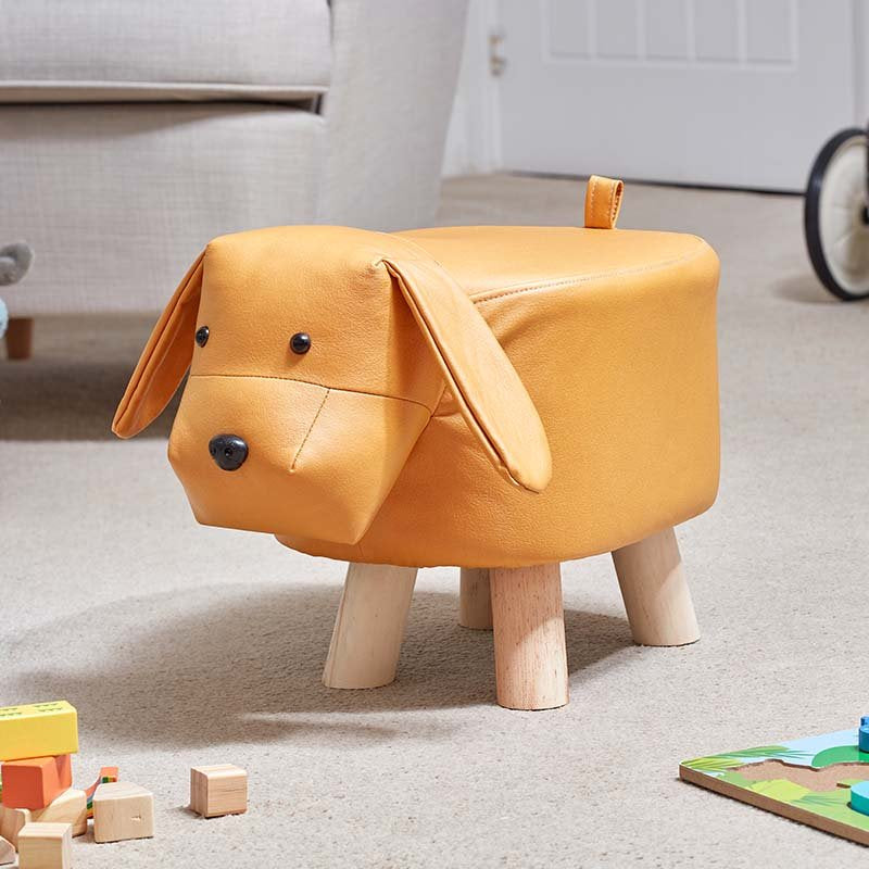 Tan leather dog-shaped footstool with floppy ears, black button eyes, and natural wooden legs, positioned on a beige carpet in a living room setting with toys nearby