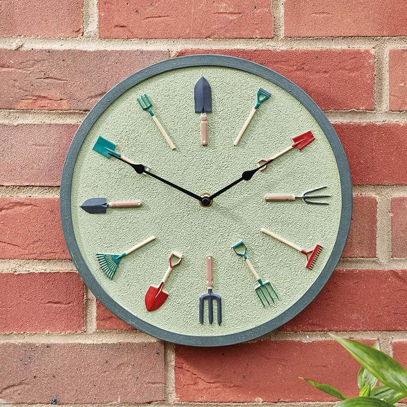 Garden tools wall clock with sage green face and gray metal rim mounted on red brick wall, featuring miniature garden tool hour markers including spade, fork, rake and trowel in teal, red and navy colors with black hands
