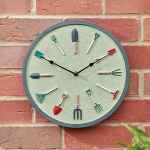 Garden tools wall clock with sage green face and metal frame mounted on red brick wall, featuring miniature gardening tool hour markers including spade, fork, rake and trowel in various colors