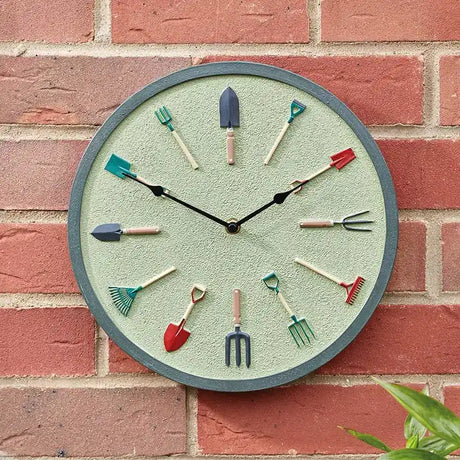 Garden tools wall clock with sage green face and metal frame mounted on red brick wall, featuring miniature gardening tool hour markers including spade, fork, rake and trowel in various colors
