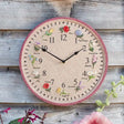 Flora Garden Wall Clock with pink border featuring embroidered flowers including rose, tulip, pansy, bluebell, lily, poppy, daffodil, lavender, daisy, gladioli, snowdrop and hydrangea positioned around clock face with black hands on cream background, displayed on wooden shiplap wall with green foliage