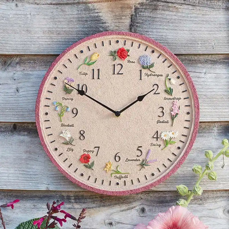 Flora Garden Wall Clock with pink border featuring embroidered flowers including rose, tulip, pansy, bluebell, lily, poppy, daffodil, lavender, daisy, gladioli, snowdrop and hydrangea positioned around clock face with black hands on cream background, displayed on wooden shiplap wall with green foliage