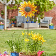 Yellow and orange metal sunflower wind spinner with black stem in garden planter surrounded by yellow lilies and pink flowers, brick house and patio furniture in background