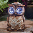 Solar-powered garden owl ornament with carved wooden design, brown and tan feather detailing, illuminated LED eyes glowing blue, perched on wooden base in outdoor garden setting