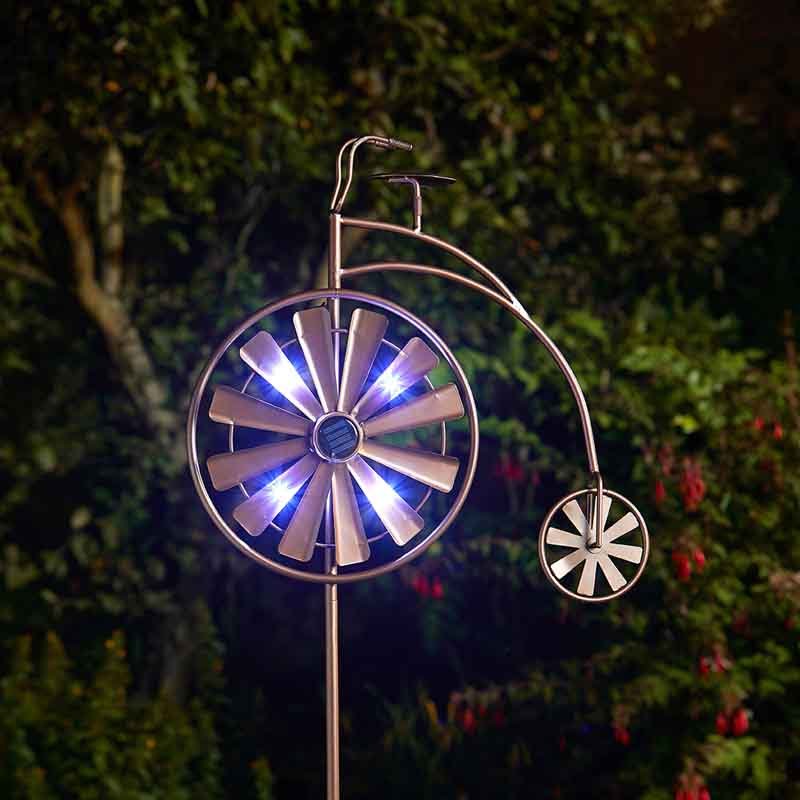 Illuminated metal penny farthing wind spinner with large glowing blue LED wheel and small decorative wheel, displayed against nighttime garden foliage with ivy and red flowers