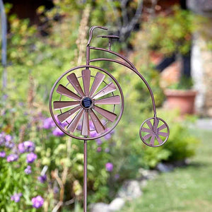 Decorative penny farthing wind spinner garden stake with purple metal wheels and frame, featuring spinning fan blades, displayed in a garden setting with purple flowers and green foliage in the background