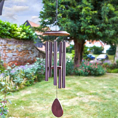 Burgundy metal wind chime with wooden top hanging in garden setting, featuring vertical tubes and teardrop clapper, displayed against green hedges and brick wall