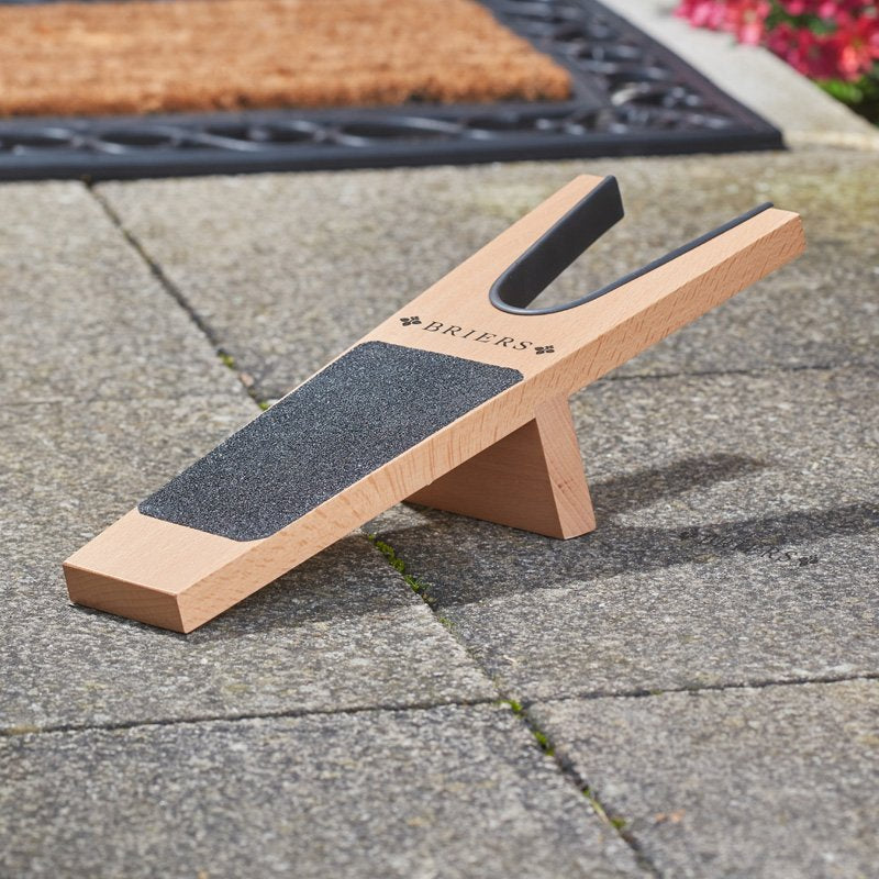 Wooden boot jack with dark gray grip pad and "Briers" branding, displayed on paved driveway with doormat and flowers in background